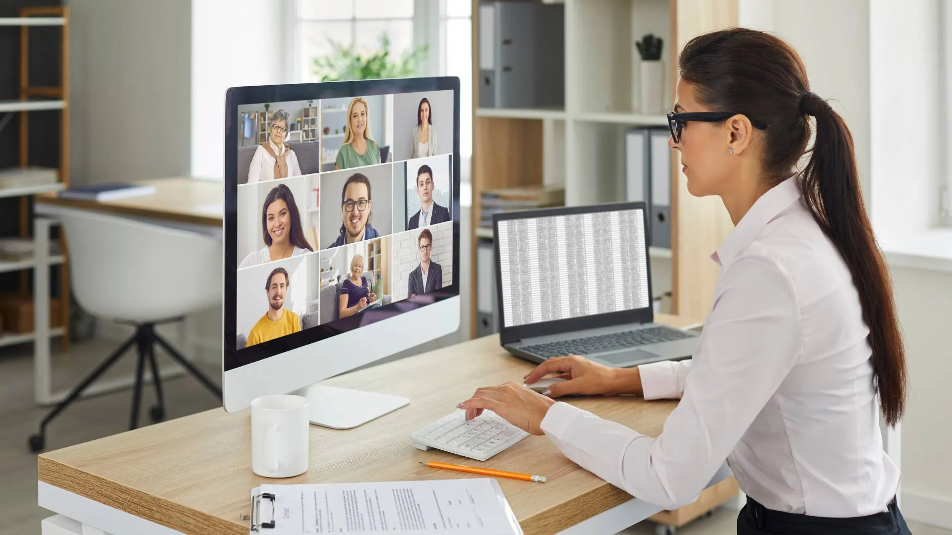Woman sitting at desk in home office in a virtual meeting on her computer. There are nine people on the screen in the virtual meeting.