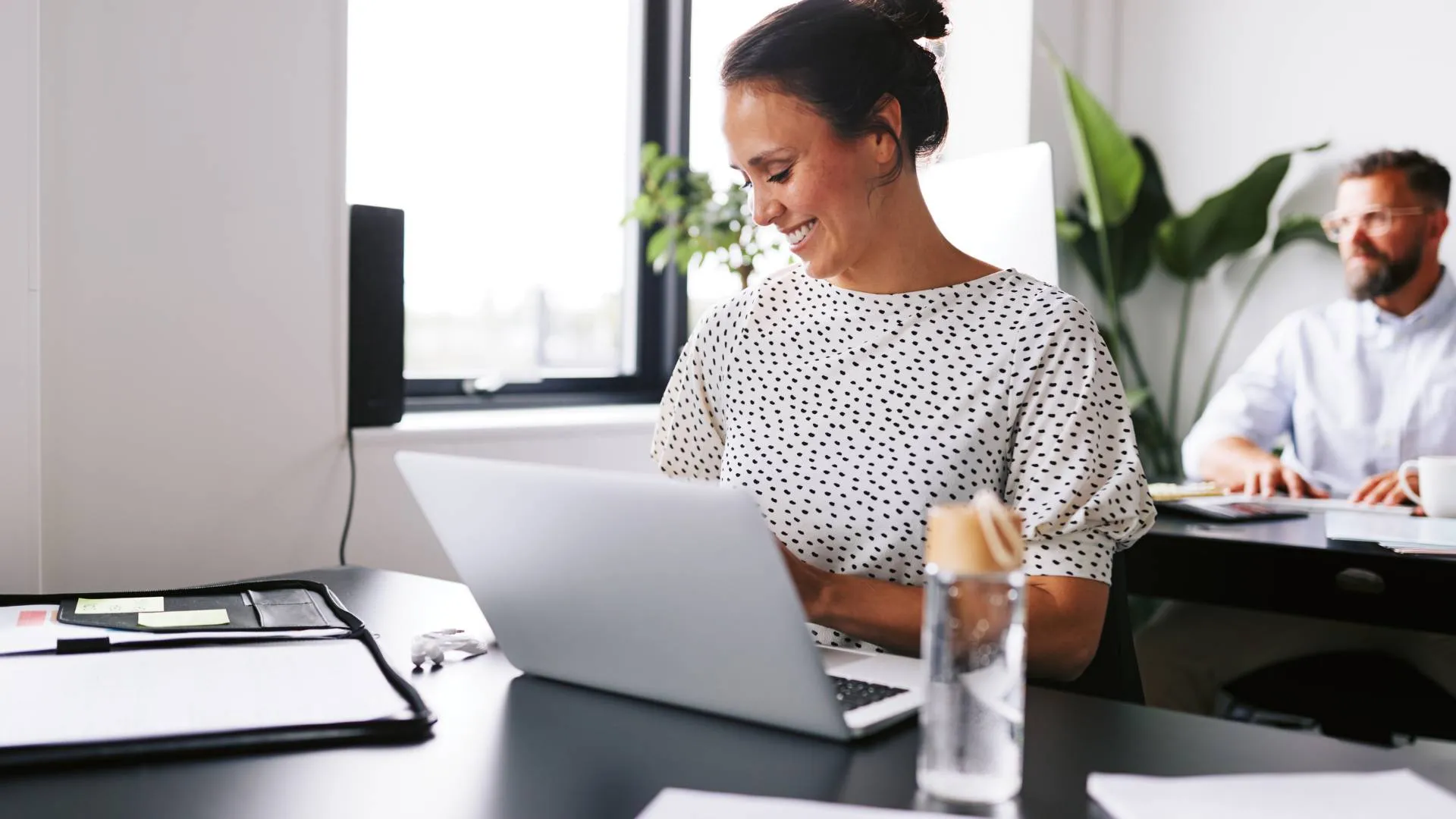 A woman who is smiling, sits at a desk in the office where she works. She's on her laptop and there's a man in the background sitting behind her at a desk on a computer.