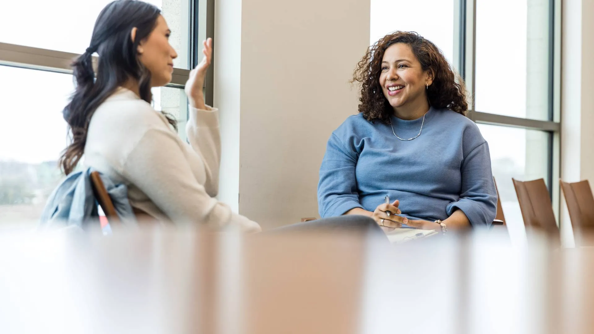 Two women sitting in an office having an engaing conversation. One woman is wearing a white shirt and the other is wearing a blue shirt.