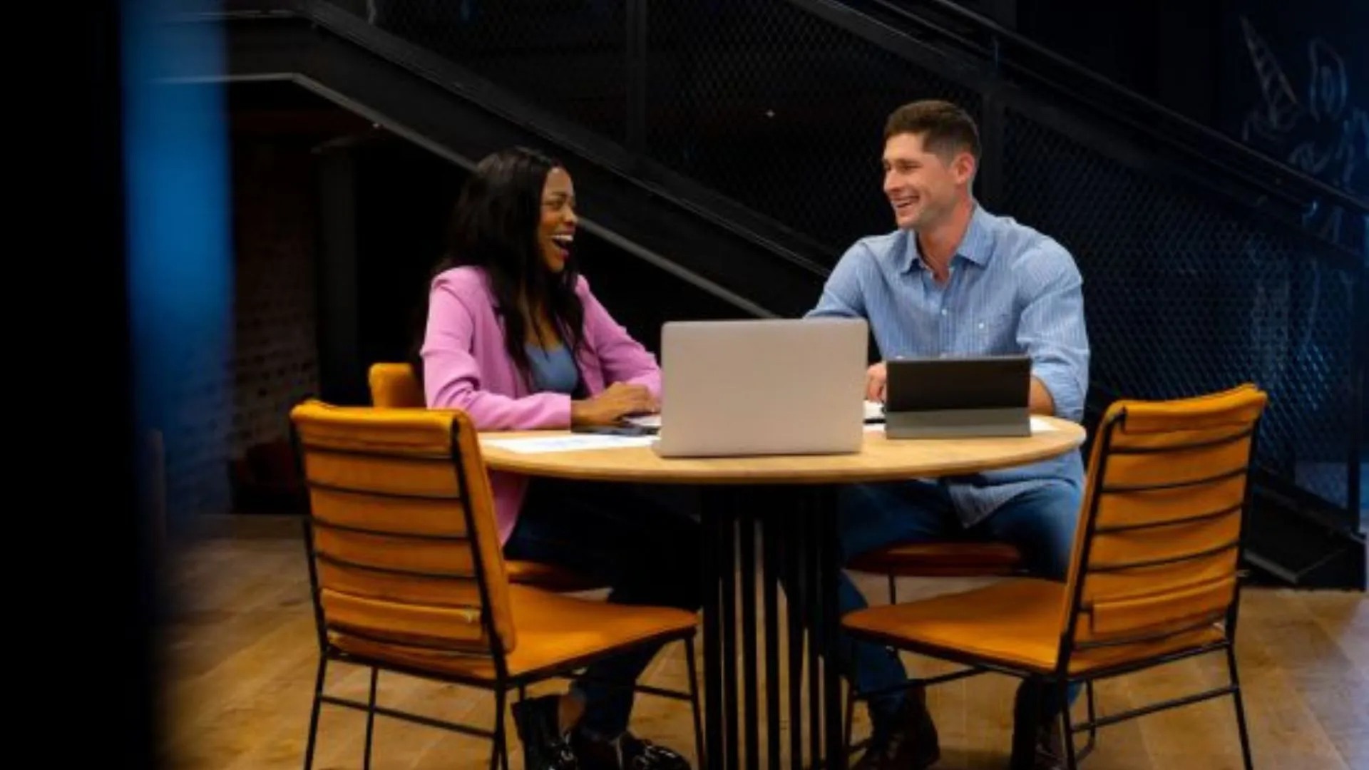 A man and woman sit at a round table during their quarterly one-on-one meeting. They’re smiling and engaging in conversation with a laptop open in front of each. They are in a casual office space with a stairway in the background, suggesting a relaxed but productive meeting environment.