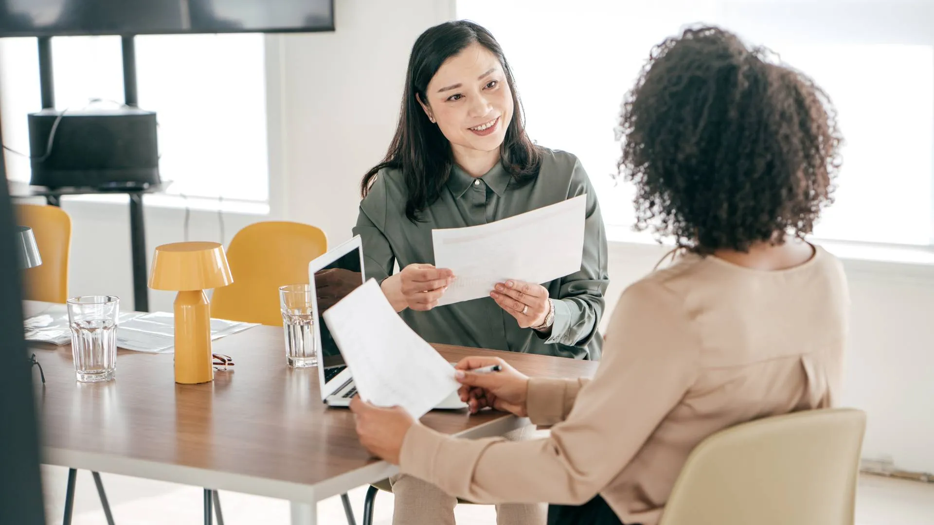 Two women are sitting at a table in an office talking. One of them is facing us while the other is facing away from us. They're going over documents they have in their hands and are sitting in front of a laptop.