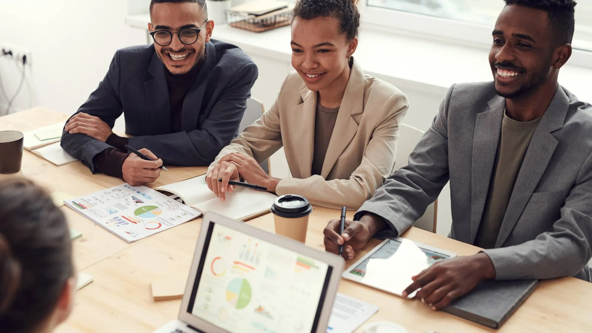 There are three employees sitting at a table together in the office engaging in a meeting with others. There's a laptop on the table with a chart displayed on the screen.