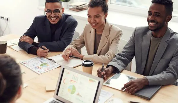 There are three employees sitting at a table together in the office engaging in a meeting with others. There's a laptop on the table with a chart displayed on the screen.