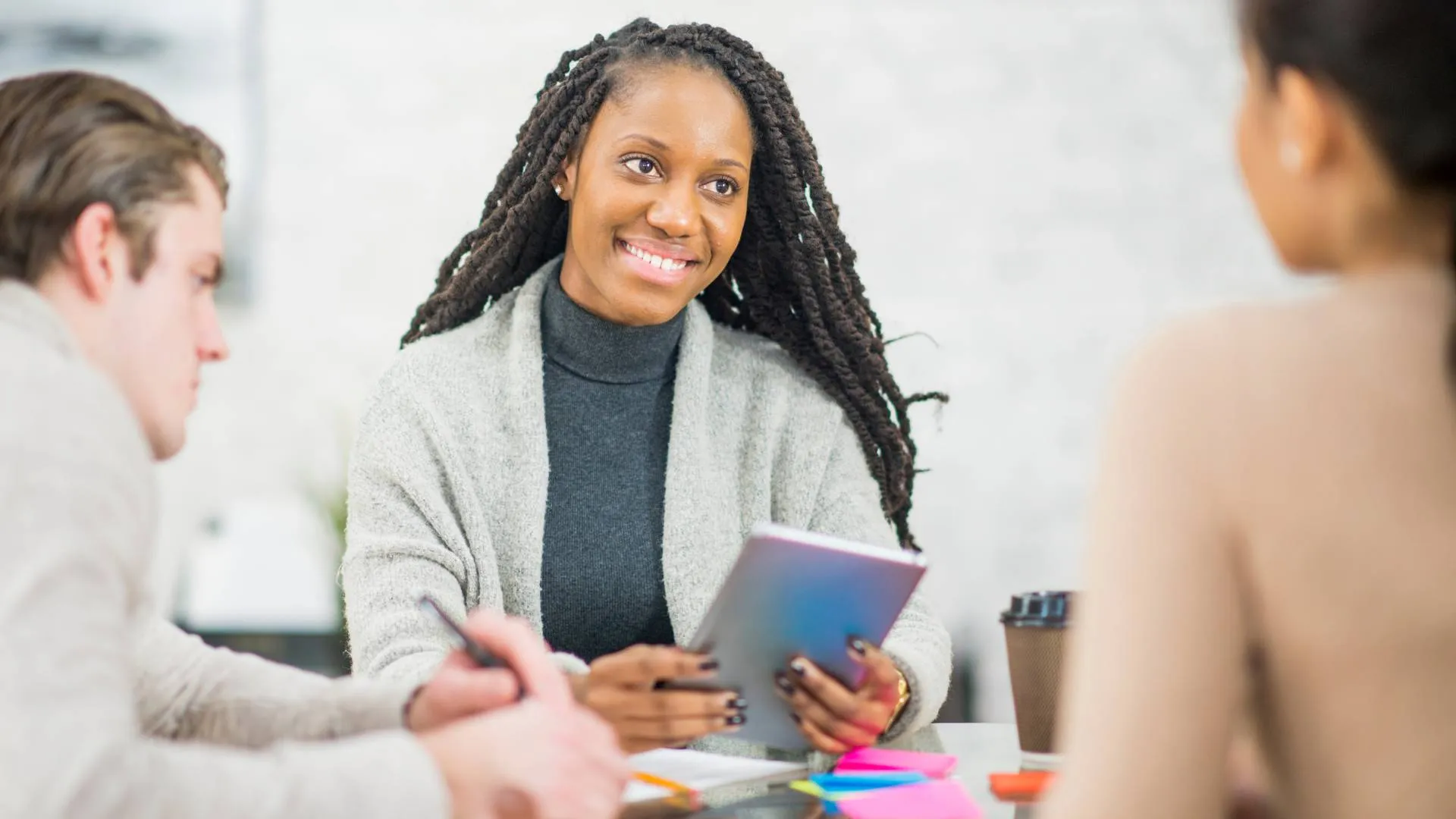 A woman is sitting at table talking to two other people who are also sitting there. She's holding a notebook in her hand.
