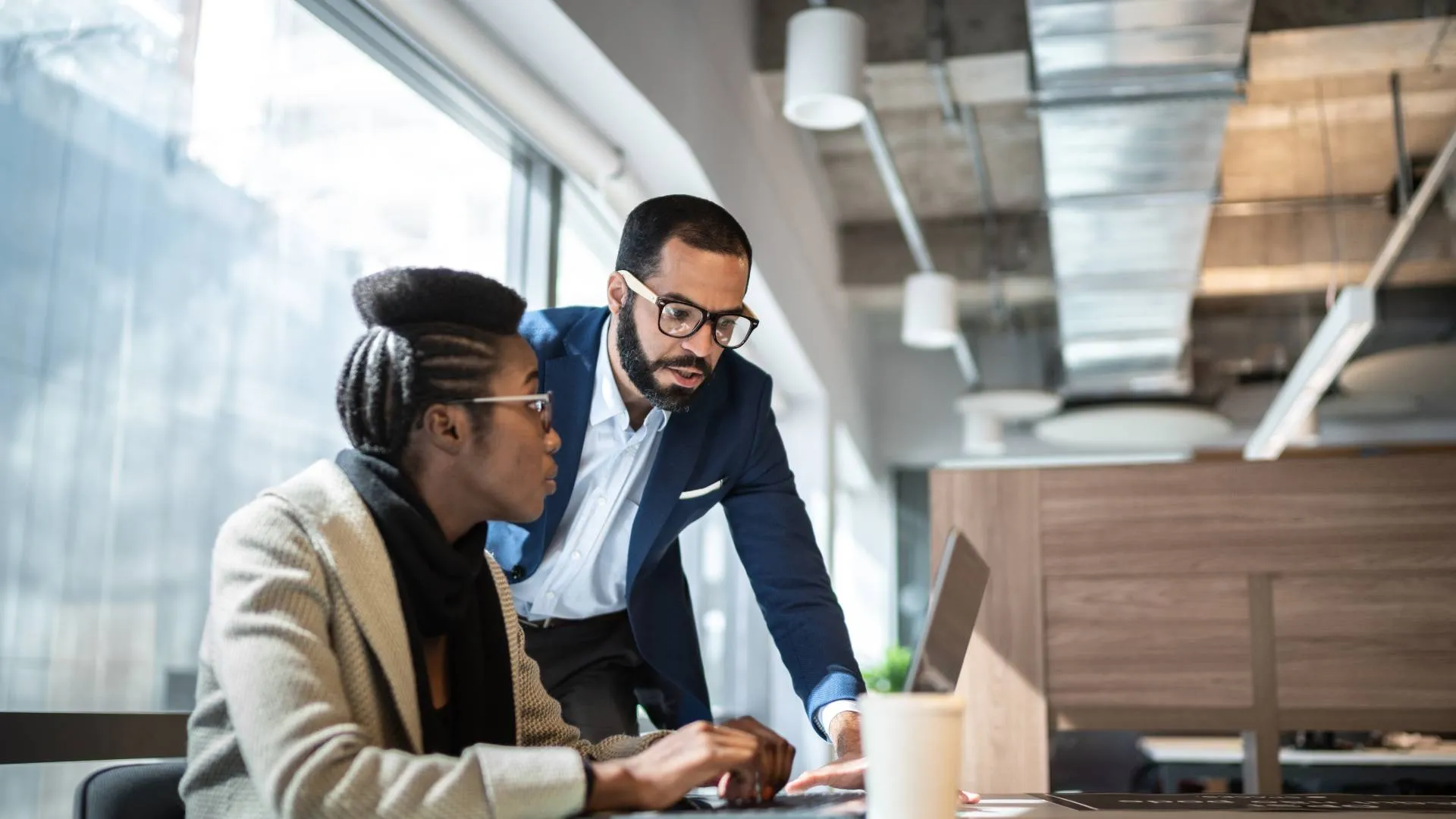 There's a woman sitting at a desk with a laptop in front of her. She's looking up at a man who is speaking to her and is standing next to her. They are both wearing glasses.