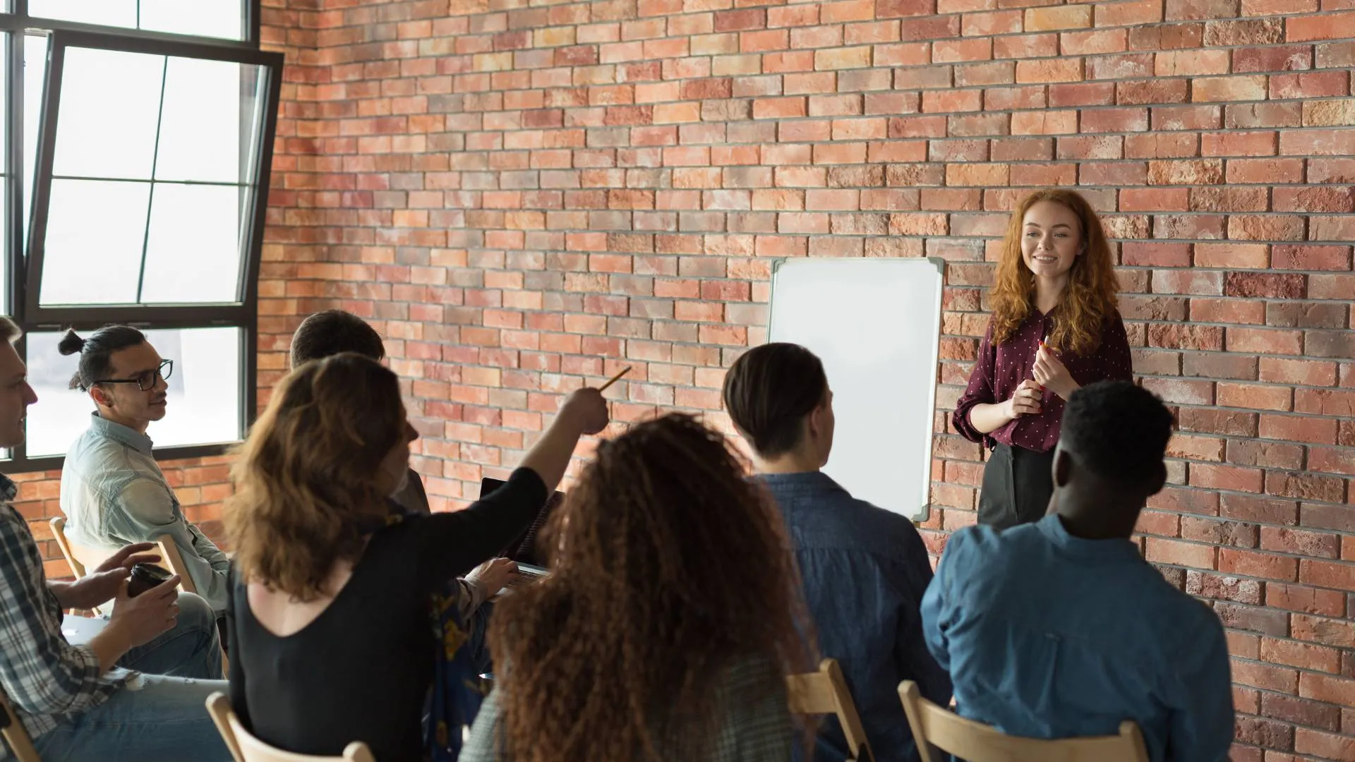 There are seven people sitting in chairs facing a brick wall where a woman is standing with a whiteboard teaching them. A woman who is seated is pointing at the whiteboard with a pencil.