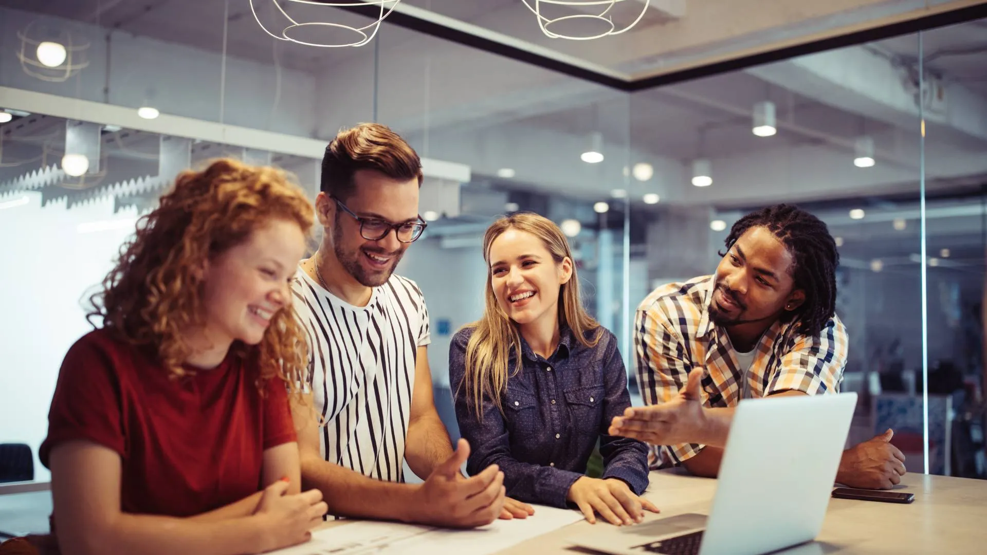 There are four people standing around a desk. They are engaged in a happy conversation and there is also a laptop on the table.