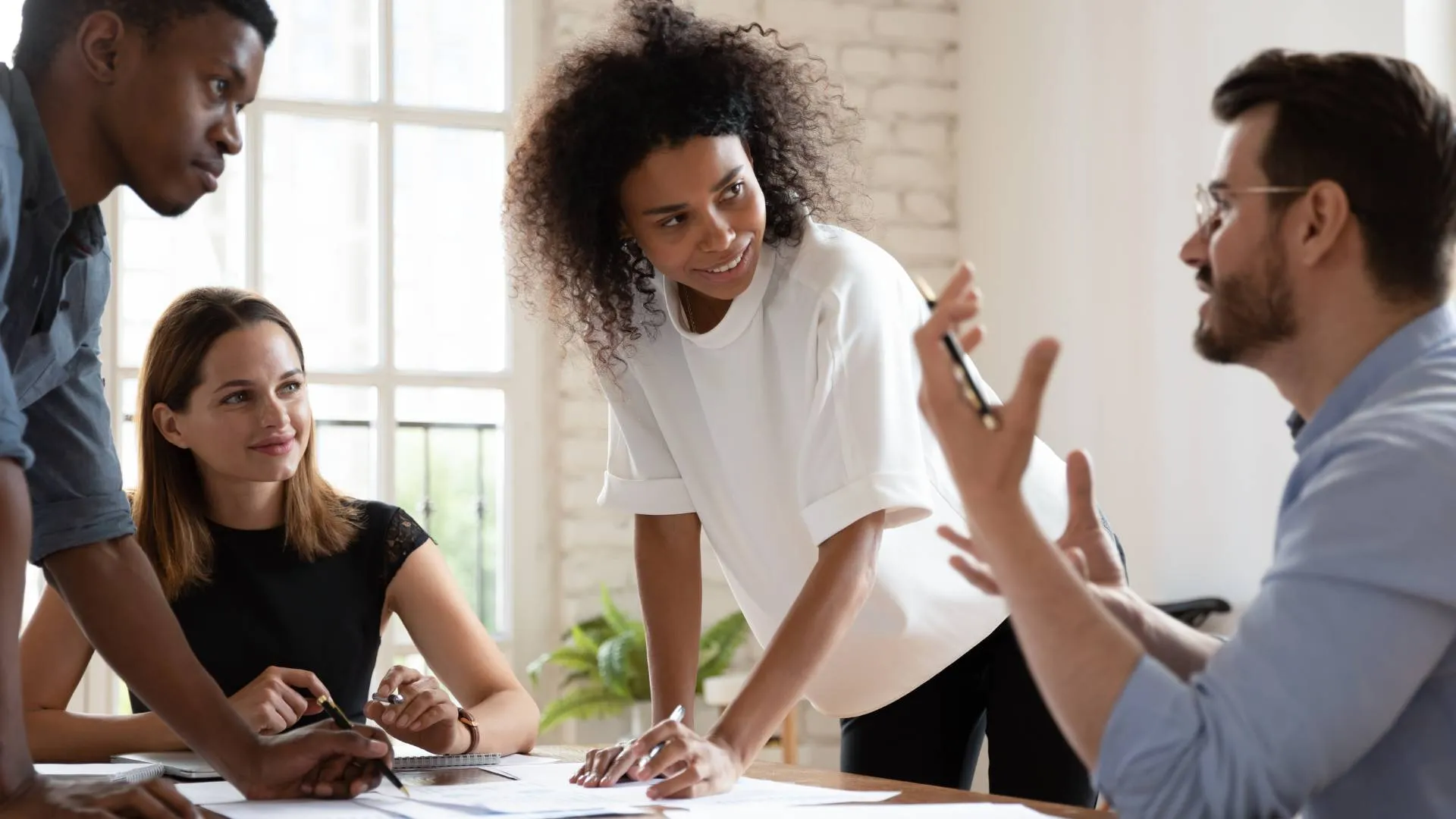 There are four people who are in a meeting. One woman is sitting down at the table along with a man who is also sitting. There is a woman and another man who are standing and engaging with the other two people.