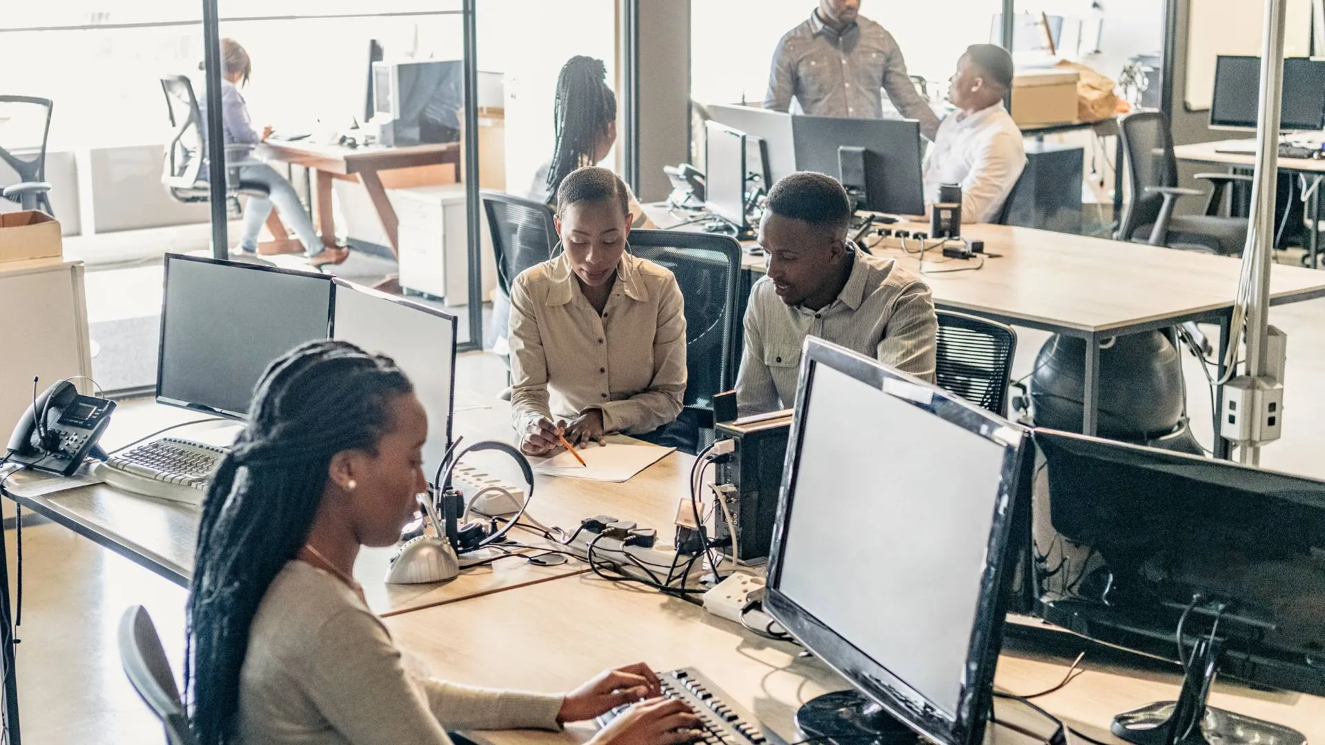 There are seven people in a large office. Three are working at a communal table which have computers on them. Another three are working at a different table and conversing. One woman is sitting by herself in another room at a desk working.