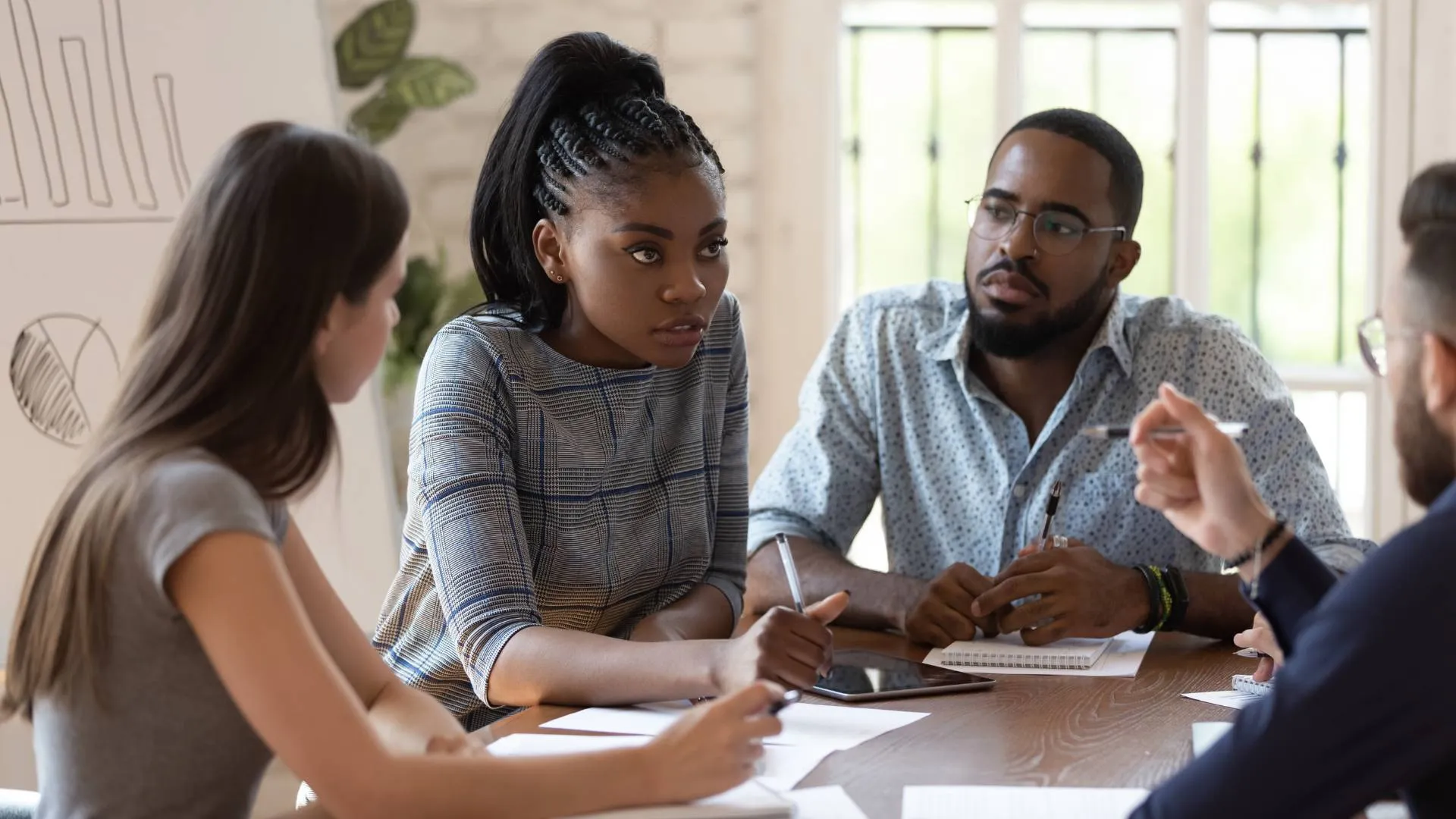 There are three employees gathered around a table in a meeting. Two are women and one is a man. There's a graph on an easel behind them.