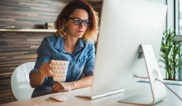 A woman with glasses is sitting at a desk holding a coffee mug. She is looking at her desktop.