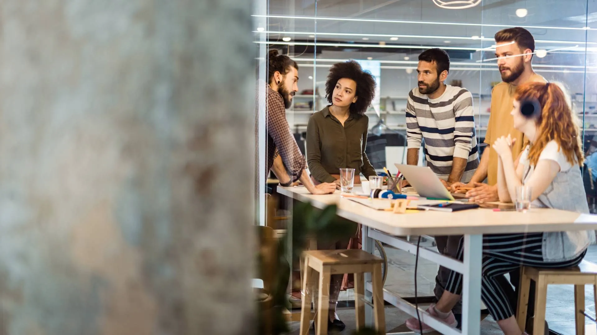 There are five team members standing around a table in an office having a meeting. One is a woman and she is sitting down and wearing a pair of black headphones. The office has an industrial feel to it.