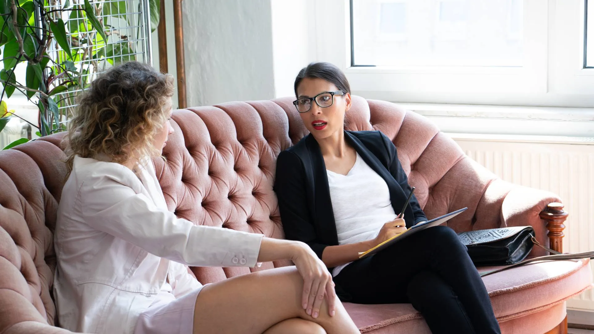 Two professional women are having a one-on-one meeting. They are sitting on a couch in an common area with plants and a window behind them.