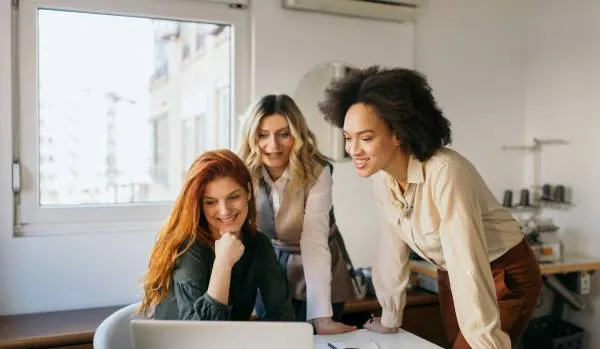 There are three women around a table smiling and looking at a laptop. They're in a meeting.