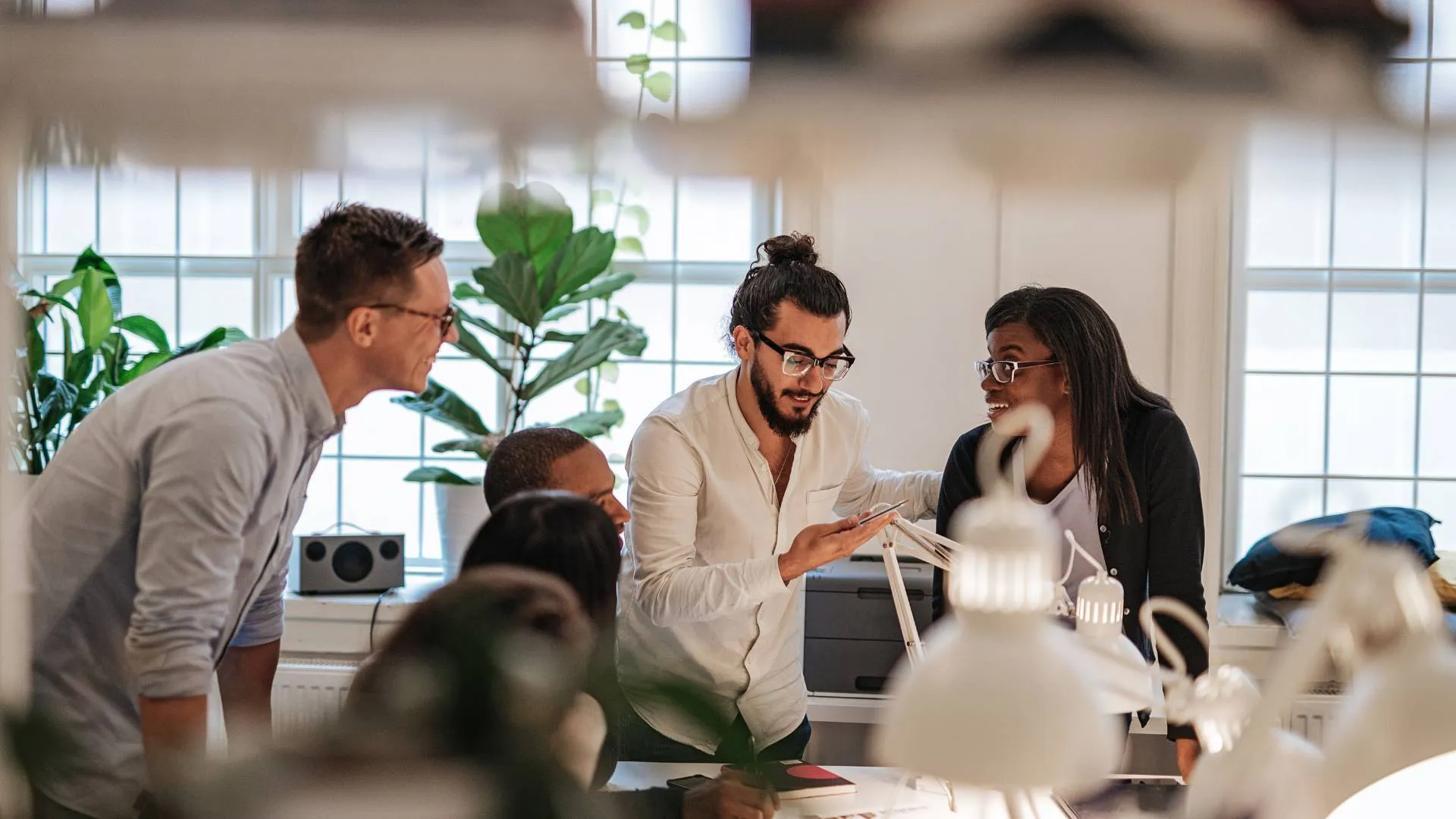 There are a group of people conversing in an office. Three are standing and the other three are sitting at a table.