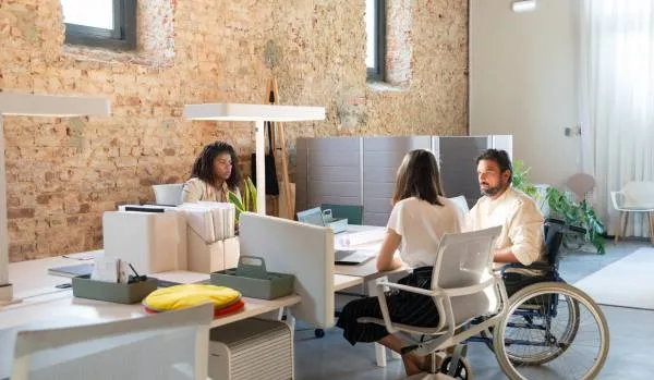 Three people sitting at a desk talking. One is in a wheelchair.