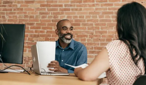 A Black man is sitting at a desk across from a woman with black hair. He is smiling while looking at her.