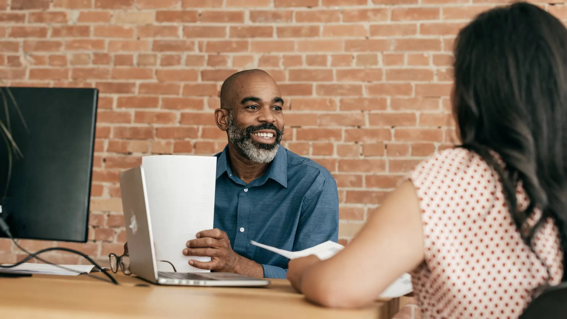 A Black man is sitting at a desk across from a woman with black hair. He is smiling while looking at her.