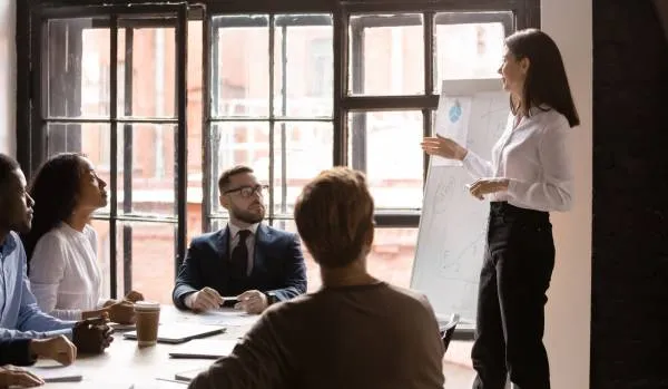 A woman who is a corporate trainer is leading a training inside of a conference room in an office setting. There are four people in the training.
