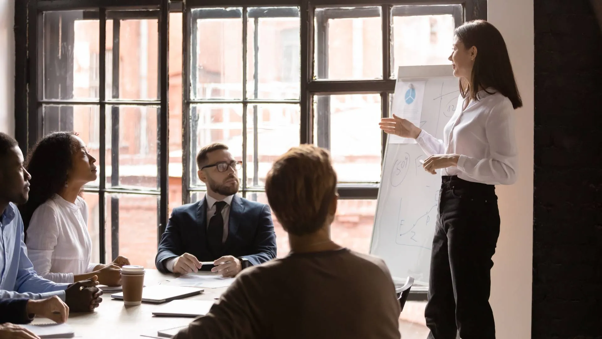 A woman who is a corporate trainer is leading a training inside of a conference room in an office setting. There are four people in the training.