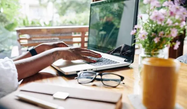 We are looking at a woman's hands typing on her laptop. She is sitting at a table on her patio with flowers, glasses, and a notebook on the table.