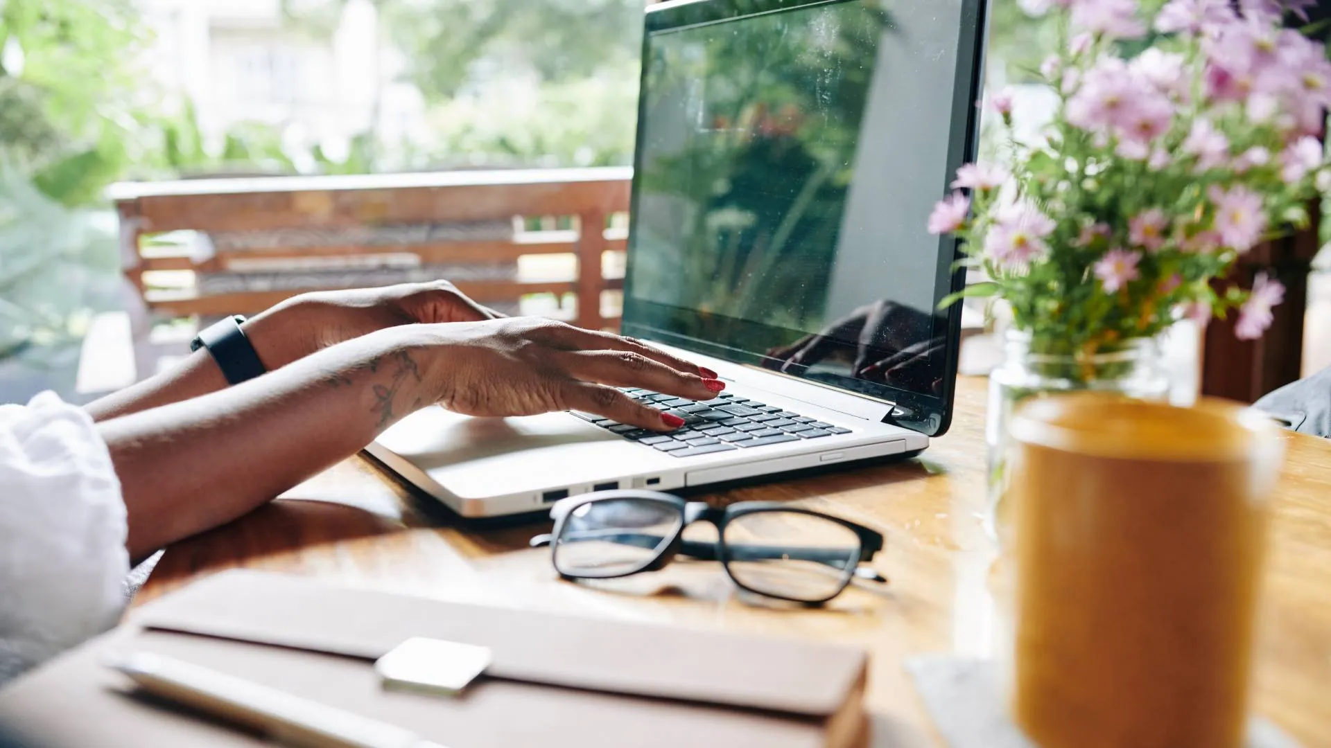 We are looking at a woman's hands typing on her laptop. She is sitting at a table on her patio with flowers, glasses, and a notebook on the table.