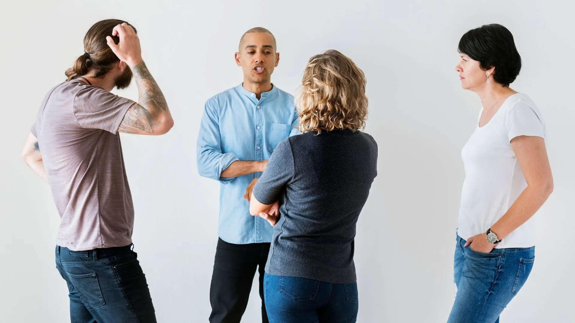 A group of four adults engaged in a discussion during a training session on healthy disagreements. The individuals, two women and two men, are in a brightly lit room with a plain white background. One man gestures while speaking to the group, one woman listens intently, another man appears stressed and is running his hand through his hair, and the other woman observes the interaction.