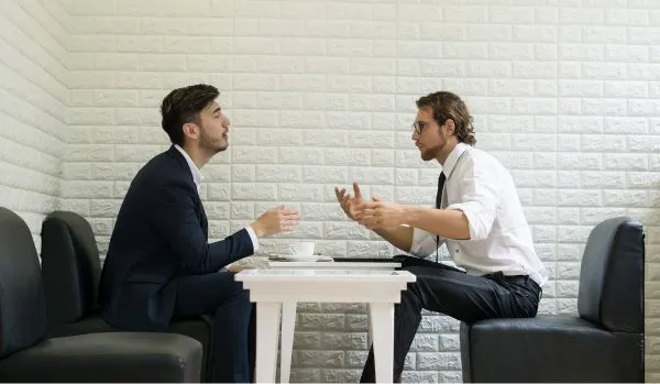 Two men sit across from a table talking during a one-on-one meeting. The manager is sitting on the left and is glad he was trained to handle emotional employees before coming to this meeting.