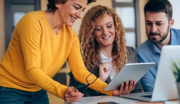 Two women and a man are smiling while looking at a tablet. The woman holding a tablet is standing next to the other two, leaning over.
