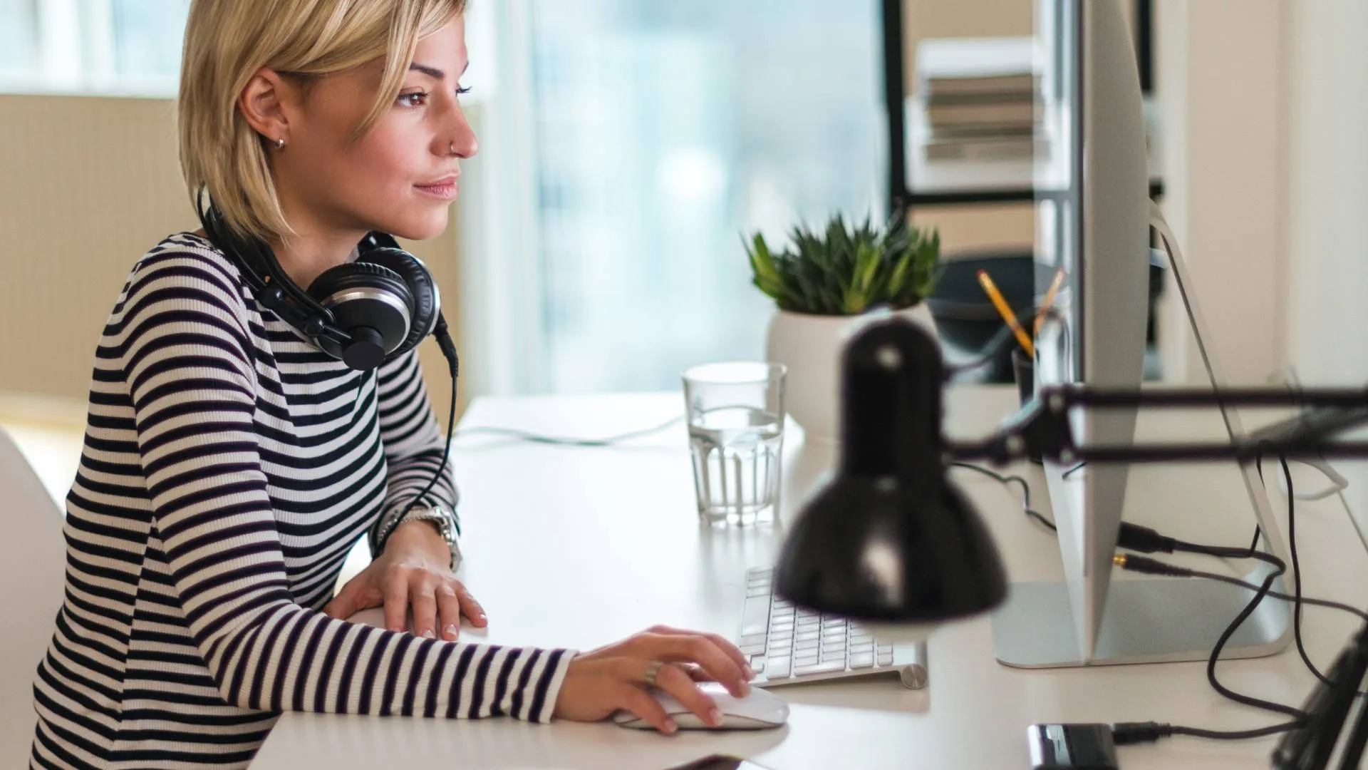 We are looking at the profile of a young woman working at her computer in her home. She has headphones around her neck.