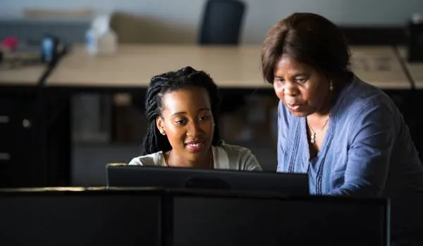 We are looking over a monitor at two woman - one standing one sitting. The older woman is standing and pointing at something on the younger woman's monitor.