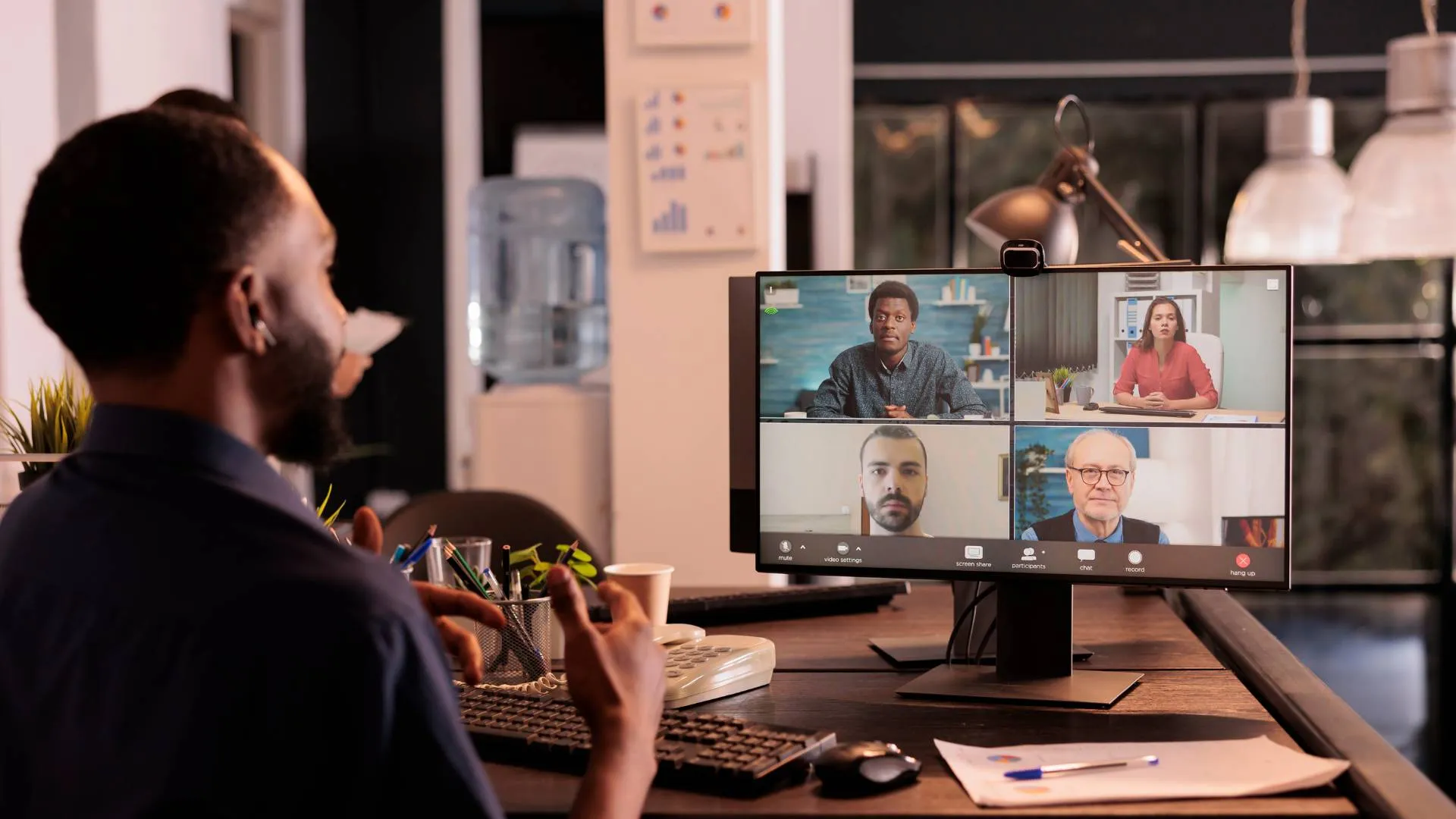 We are looking over a man's shoulder at his computer monitor on his desk while he talks to his team on video conference.