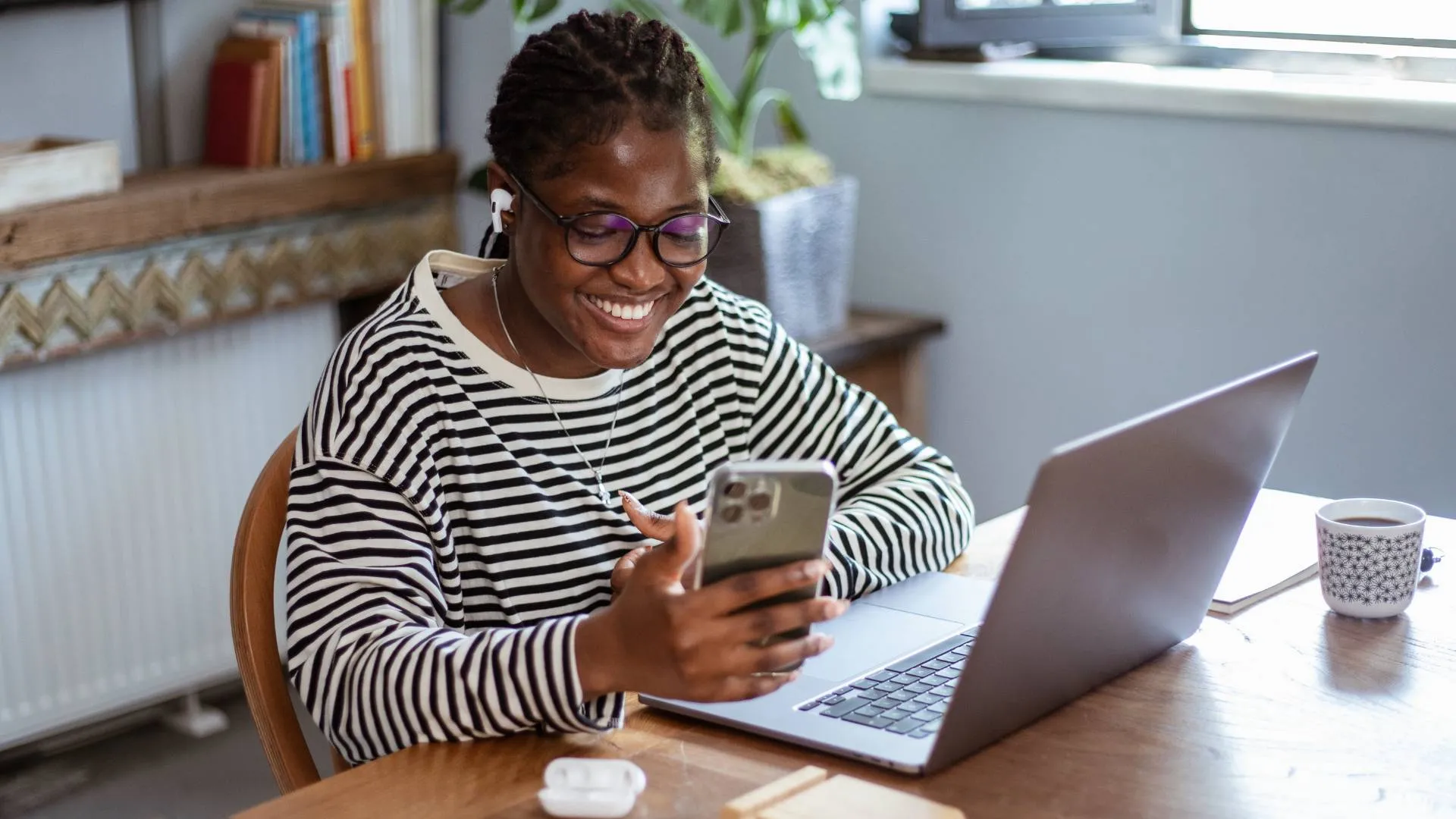 A young woman is working at her laptop on her desk, smiling and looking at her phone.