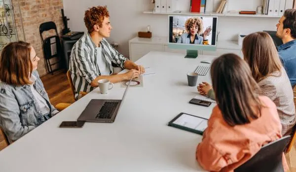 A team of people are seated around a conference table looking at a colleague on-screen at the end of the table. 