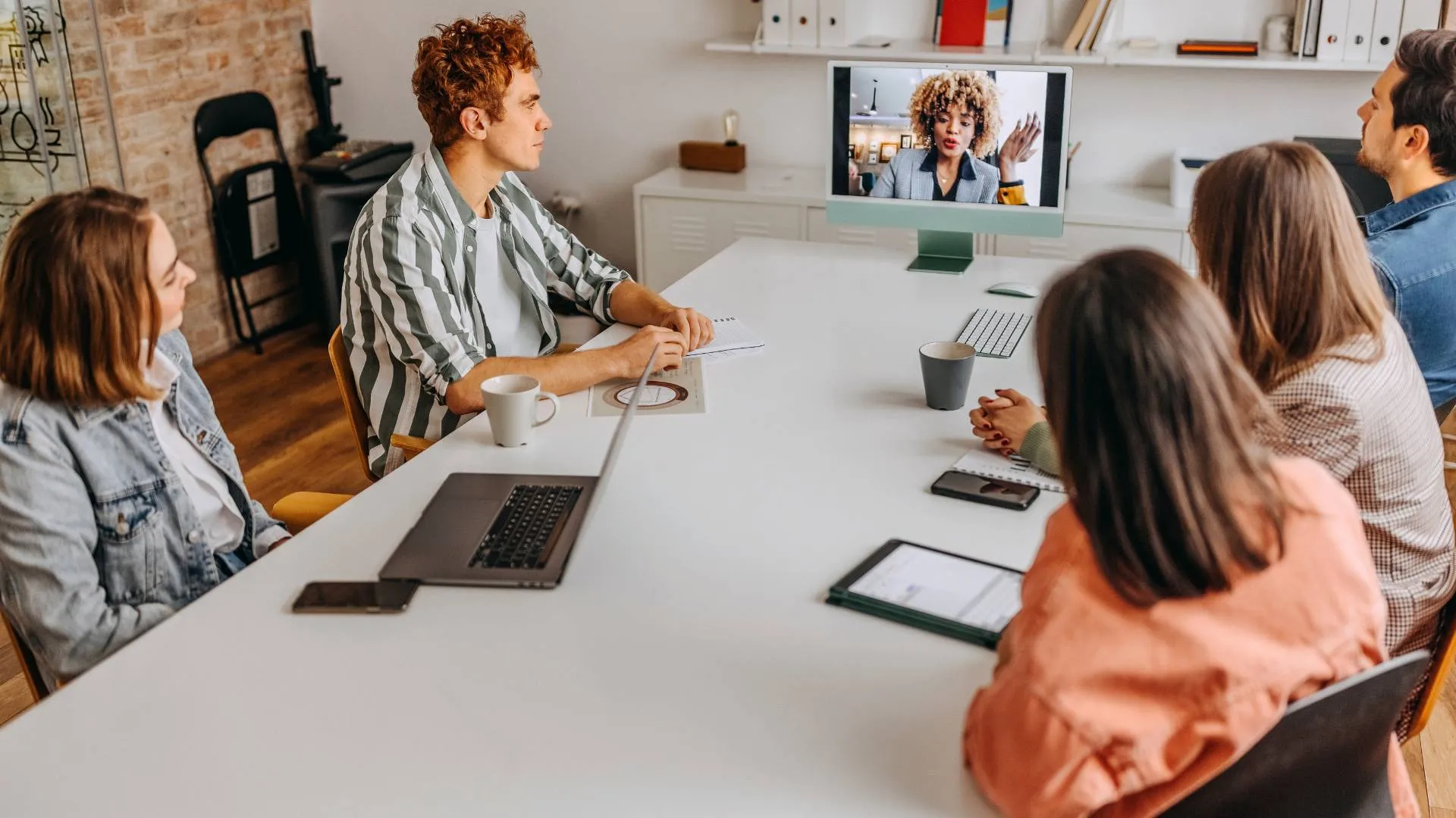 A team of people are seated around a conference table looking at a colleague on-screen at the end of the table. 