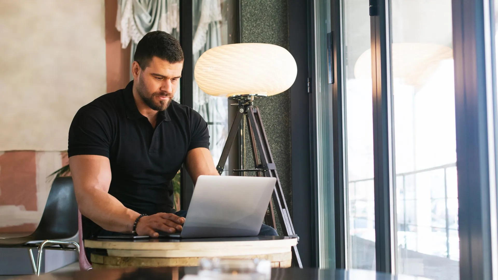A man is working on his laptop at a small table in a common area near a window.