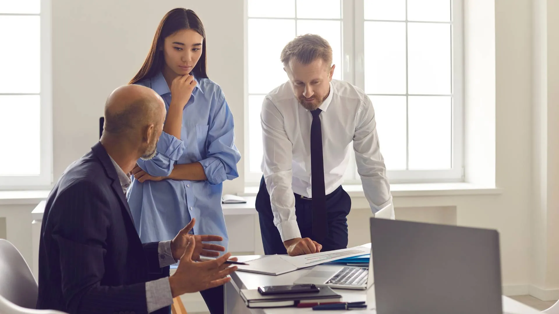 A boss is sitting at his desk having a conversation with two younger employees who are standing and showing him some documents.
