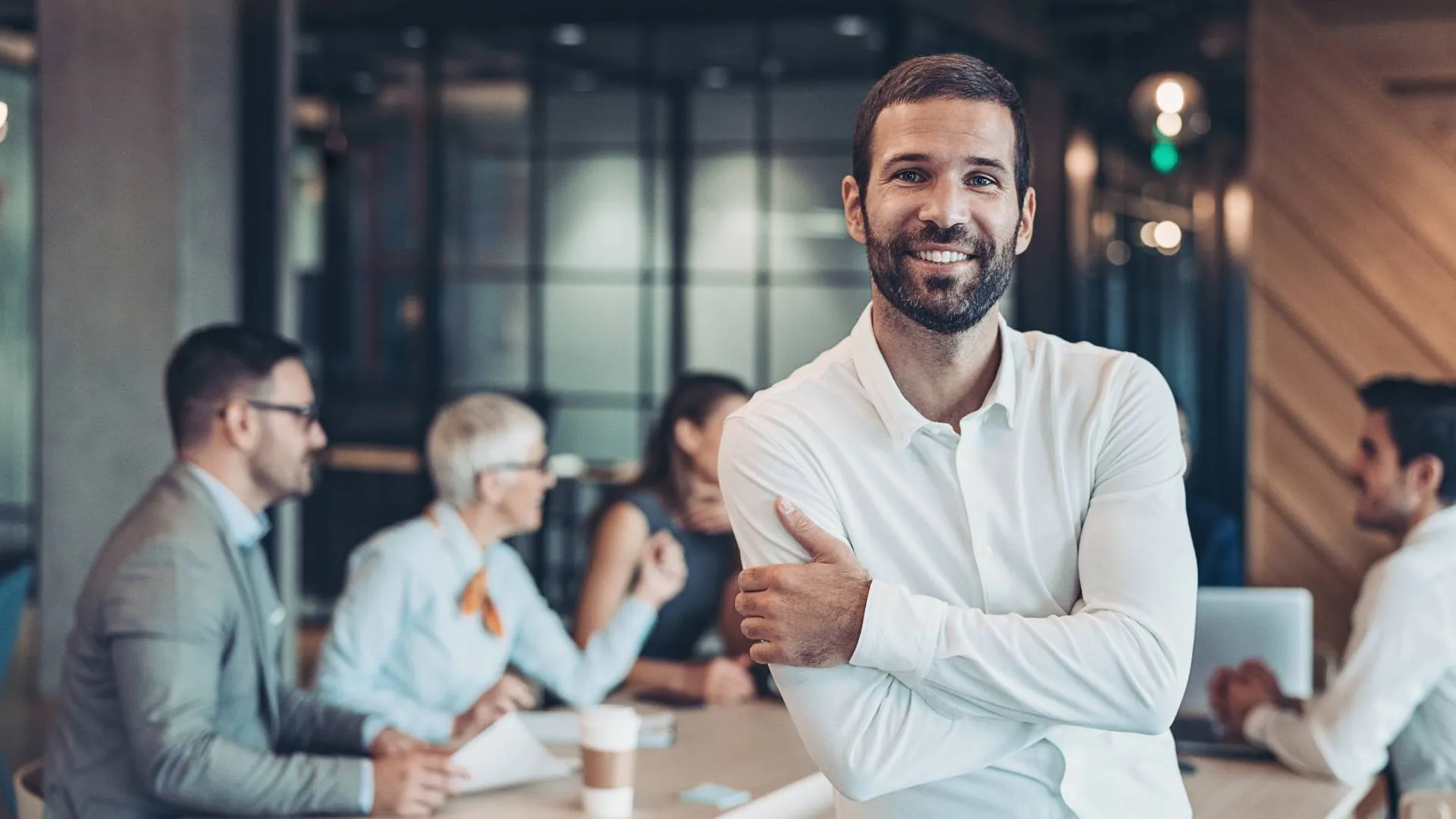 A man is in the foreground smiling at the camera. Behind him, his team works together at a conference table.
