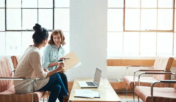 Two women are sitting in an open common area discussing a project. One is holding a folder with papers and the other is holding a coffee.