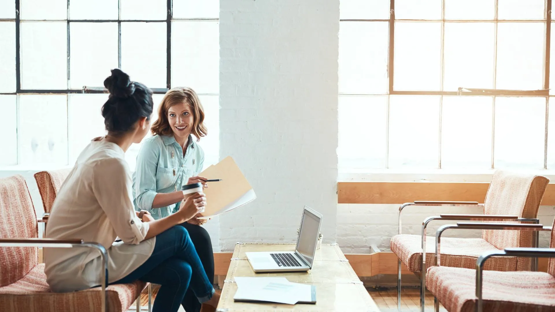 Two women are sitting in an open common area discussing a project. One is holding a folder with papers and the other is holding a coffee.