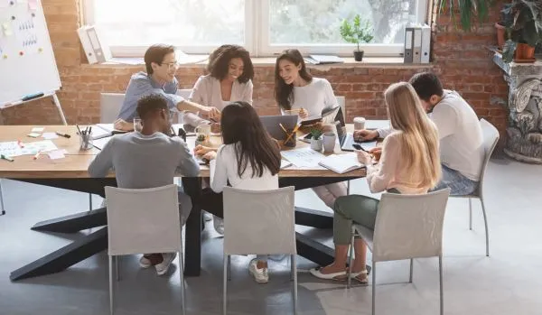 A diverse group of employees are working together on a project around a conference table with a window in the background.