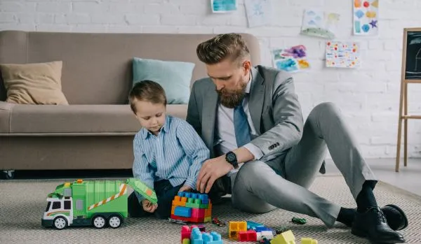 A man in a suit is sitting on the floor of his living room playing with his child.