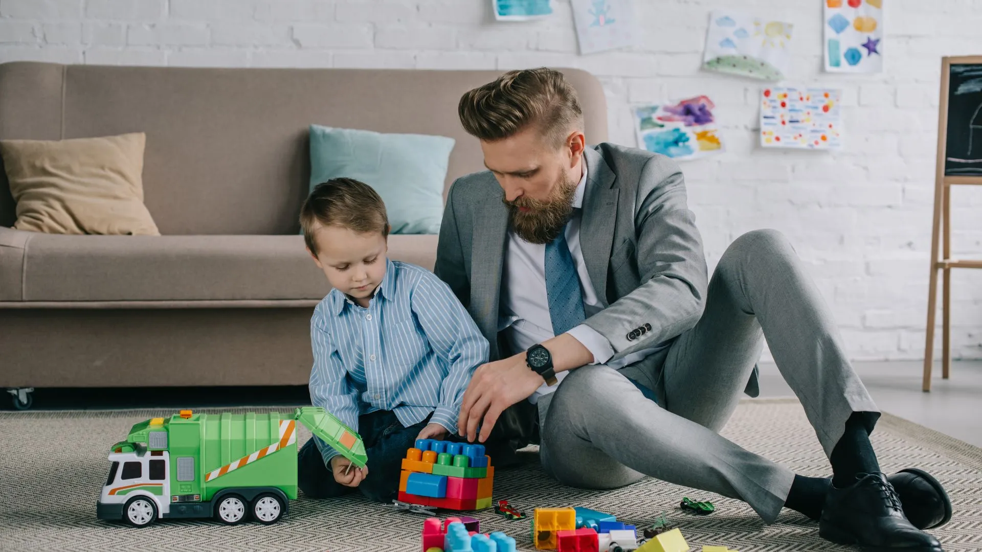 A man in a suit is sitting on the floor of his living room playing with his child.