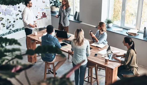 A diverse team of people are standing and sitting around a tall conference table looking at two men in front of a flip chart during a brainstorming session.