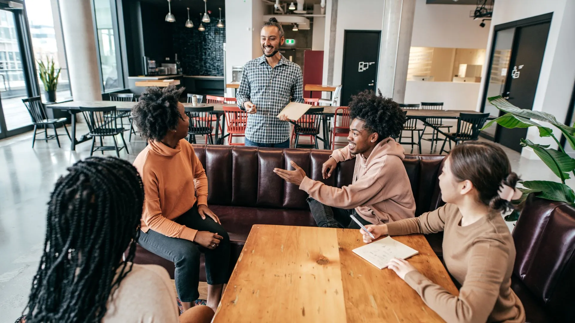 A man with a bun and a beard is standing in front of a table of diverse young managers training them.