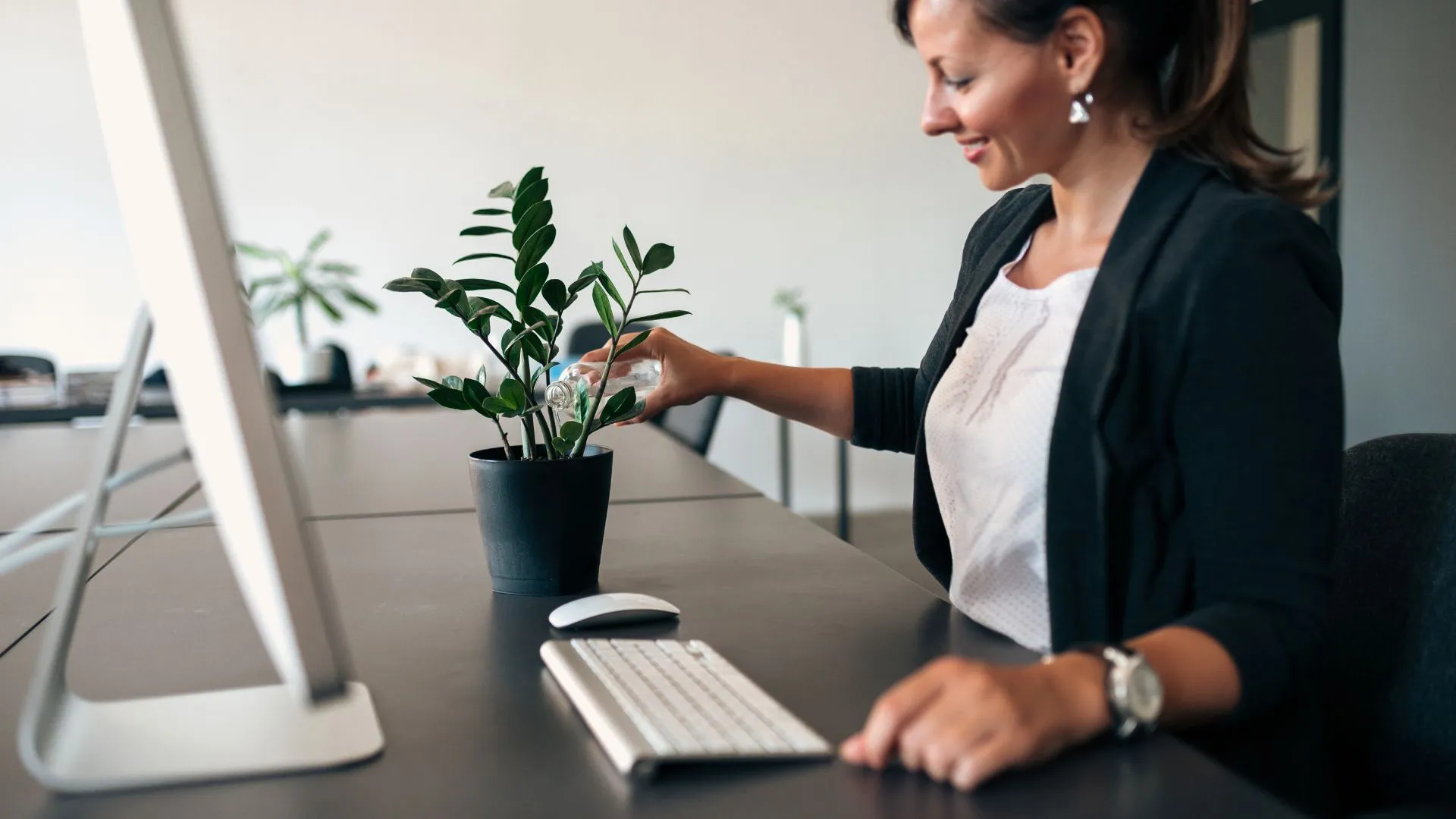 A woman is watering a plan on her desk. We are looking at her from the side.
