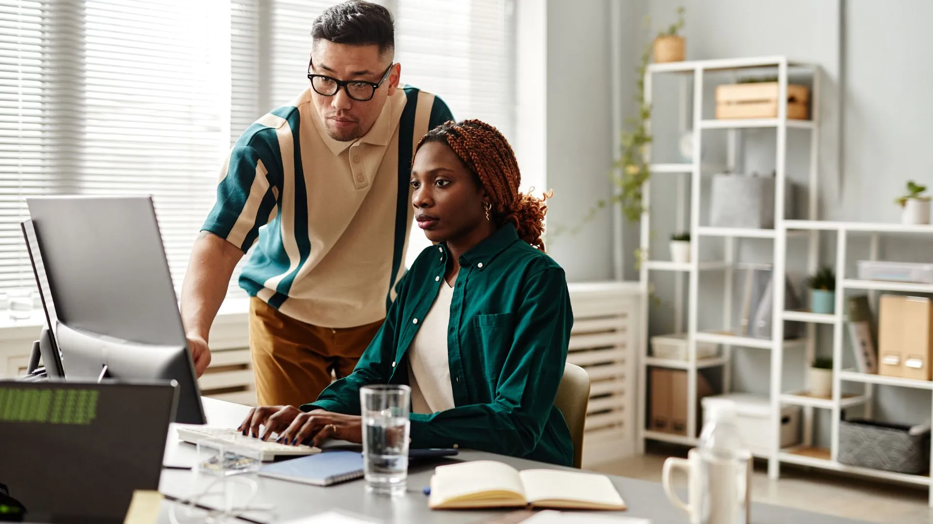 A man is standing behind a woman at a desk, pointing at something on her computer.