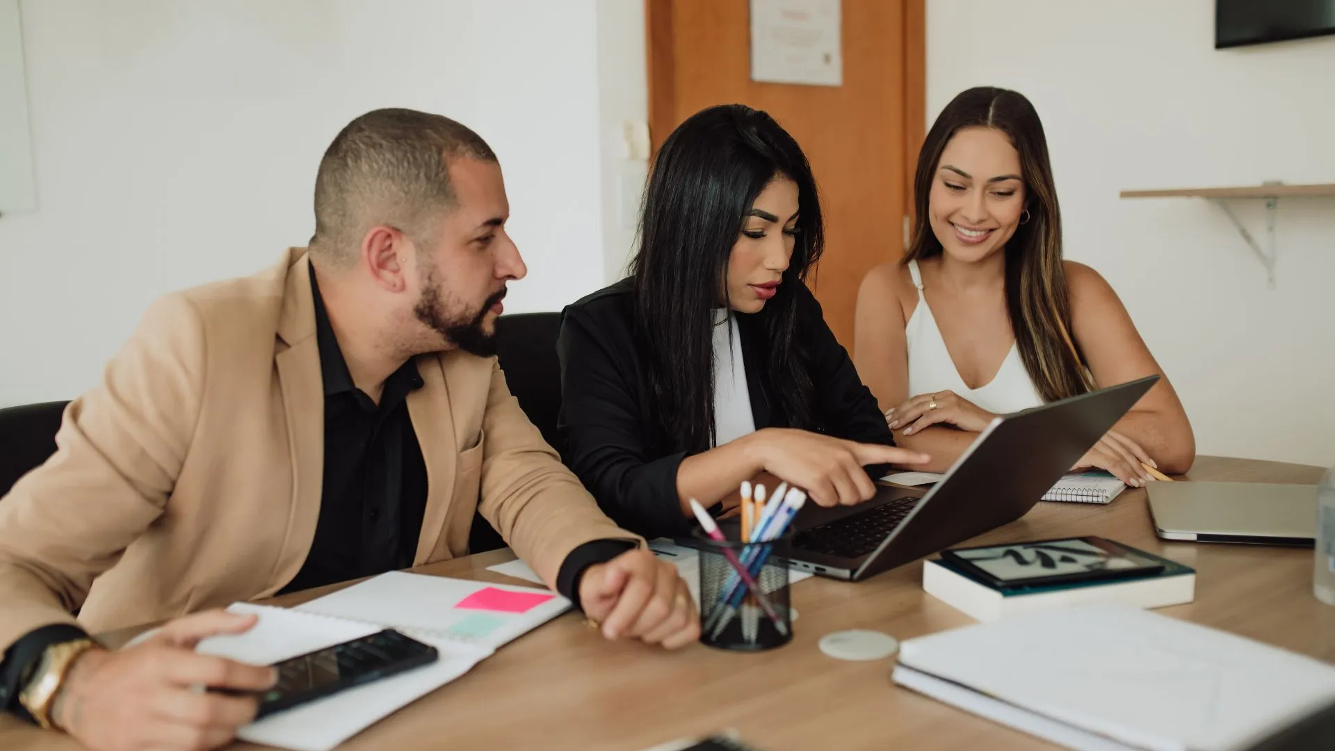 Three people are working together at a conference table. Two of them are looking over at the middle person's laptop.