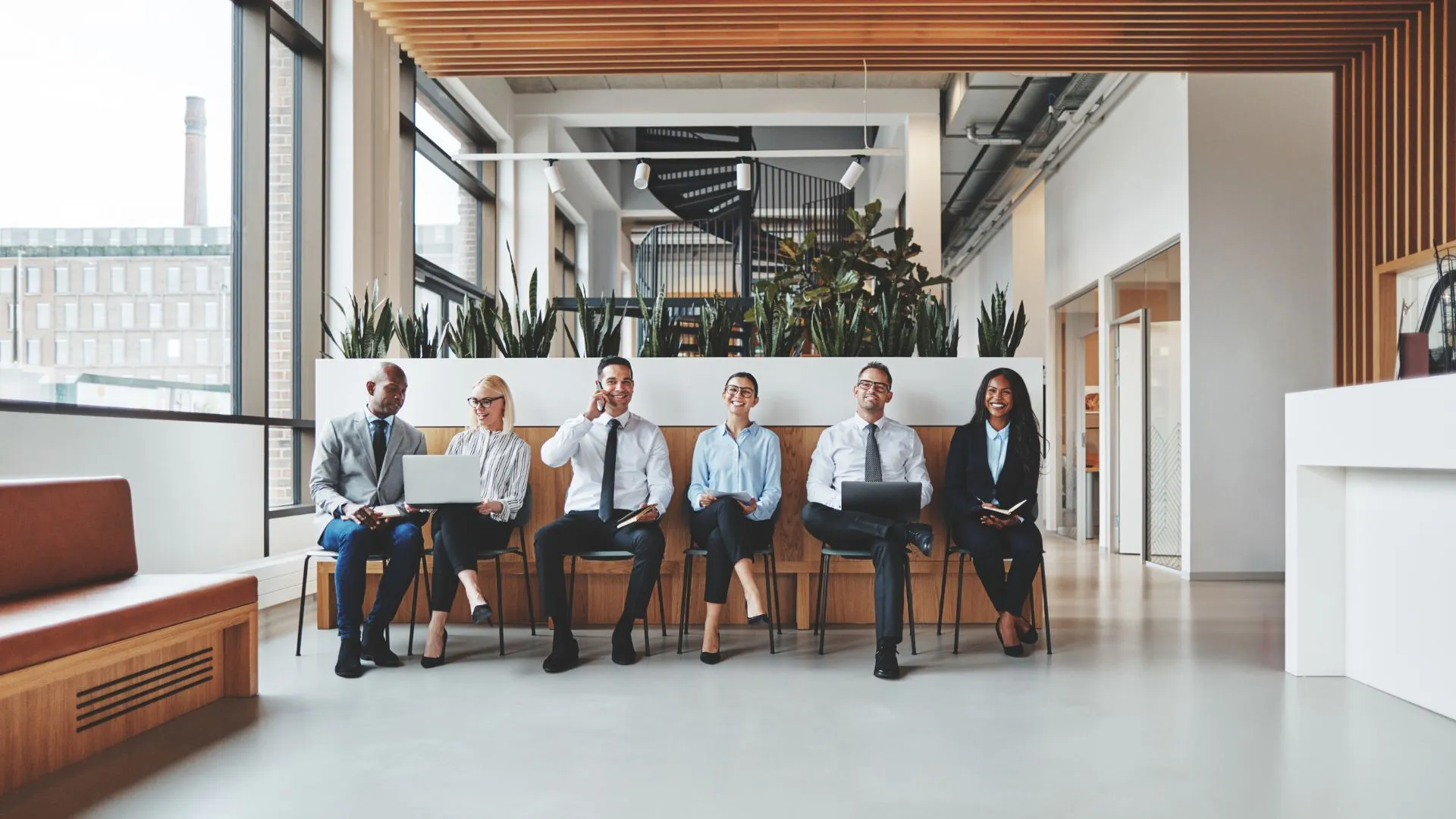 A diverse group of job applicants are sitting in a row in the lobby of a modern office. They are laughing and conversing.