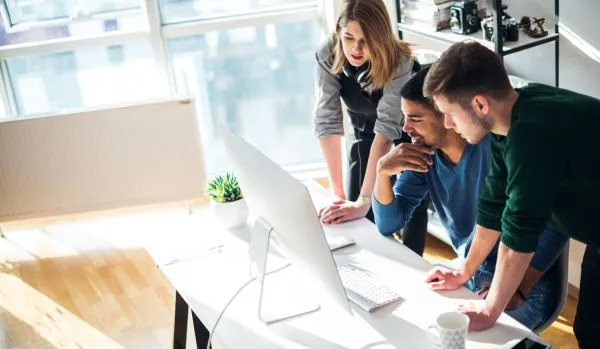 A group of three people are gathered behind one person's desk looking at that person's computer monitor.